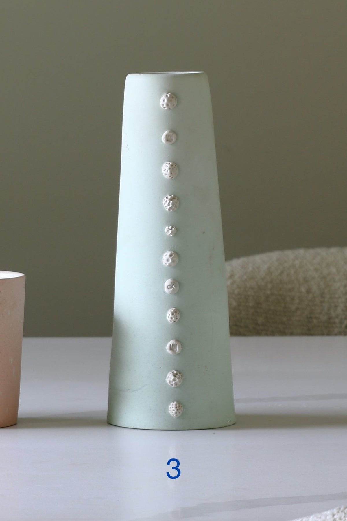 A tall, tapered ceramic vase in a pale mint green glaze, decorated with a vertical row of textured, raised white circular accents. The vase is displayed on a light-colored tabletop with a neutral background.