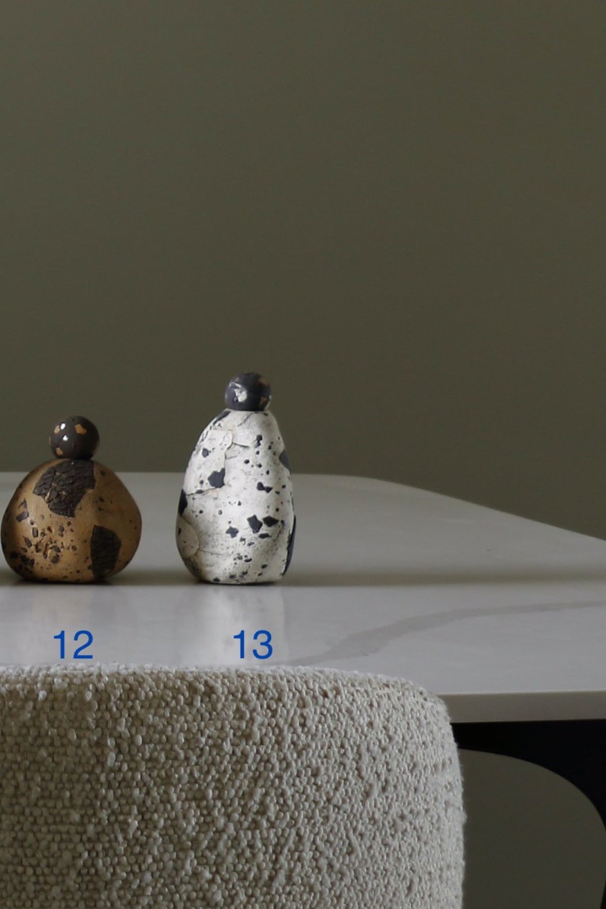 Two small decorative bottle-shaped objects displayed on a white tabletop against a muted olive-gray wall. The left object is brown with dark patches and the number 12 below it; the right object is white with black speckles and the number 13 below it.