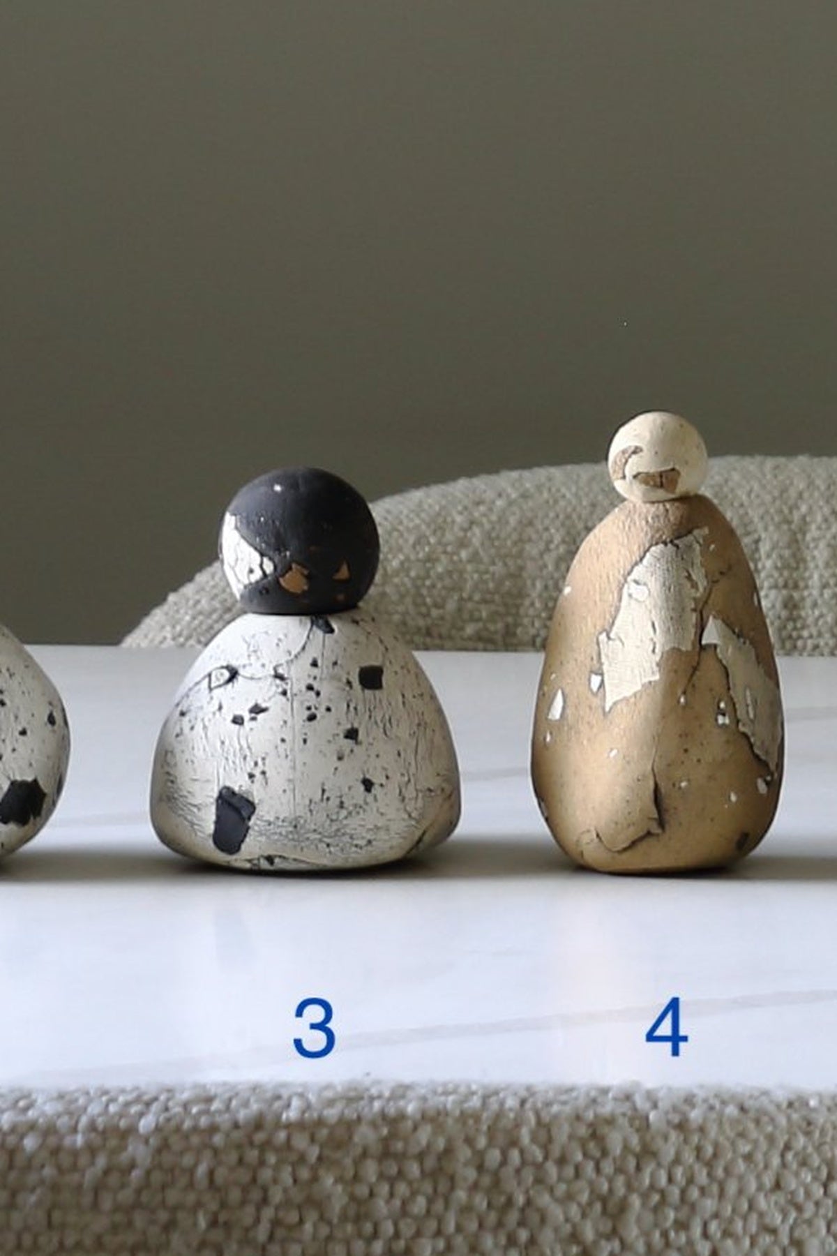 Two decorative ceramic figurines with a distressed, peeling glaze finish are displayed on a white marble surface. The left figurine is off-white with black speckles and a black top, labeled with the number 3.