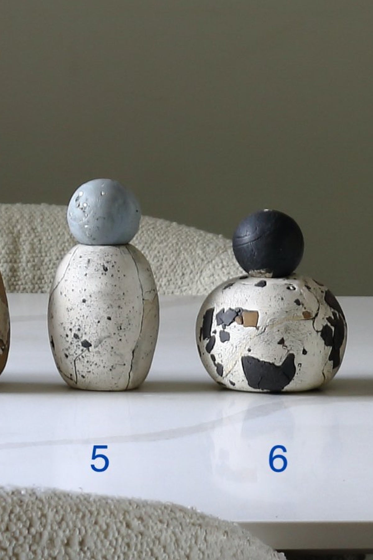 Two decorative ceramic figurines with textured, speckled painted surfaces are displayed on a white surface against a neutral background. The left figurine has a light blue spherical top and the right figurine has a dark blue top.