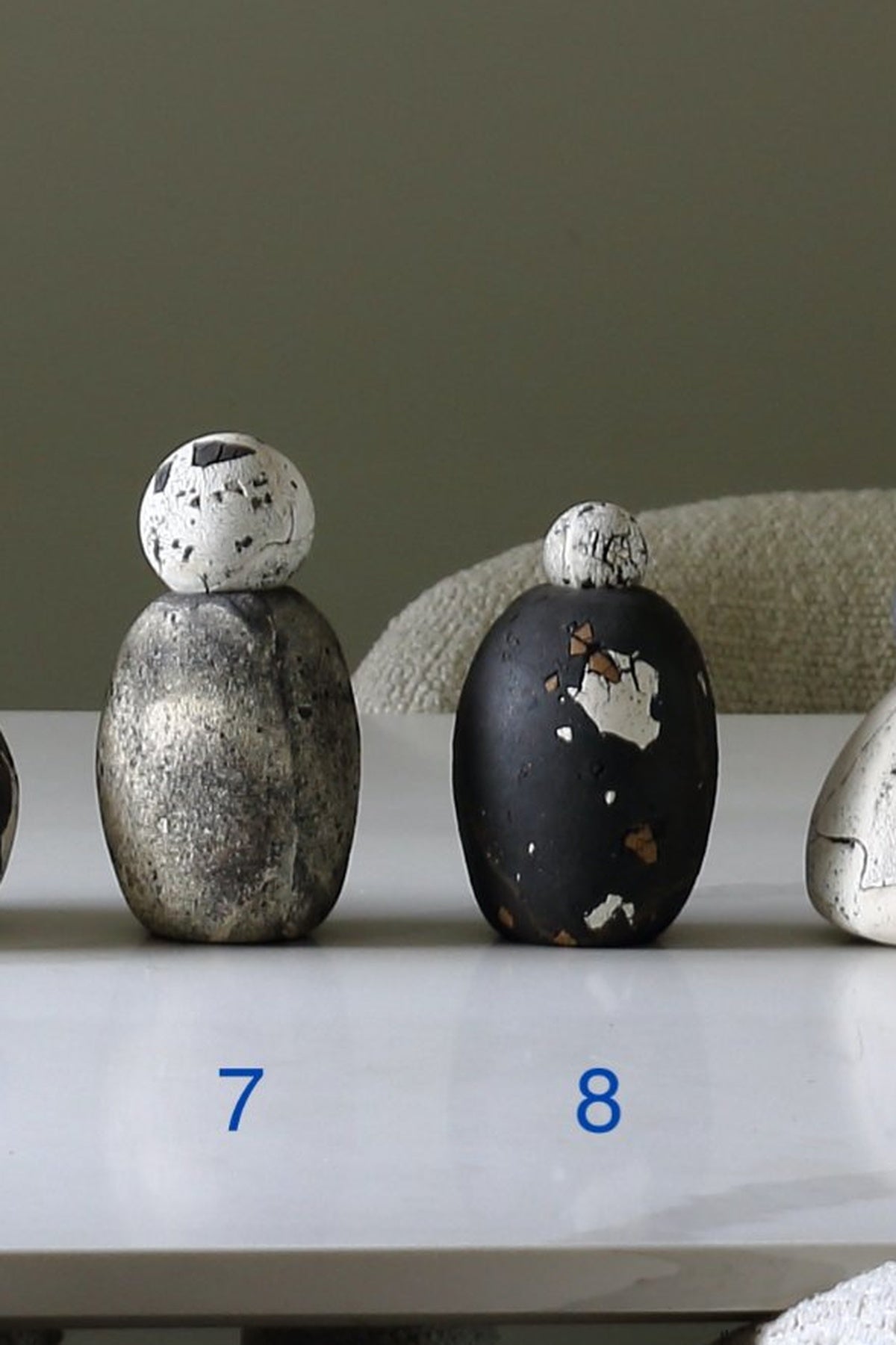 Two small decorative stone-like vases or figurines displayed on a white surface against a muted gray-green background. The left piece is metallic silver with a white speckled top, and the right piece is black with white and brown marbling and a white top.
