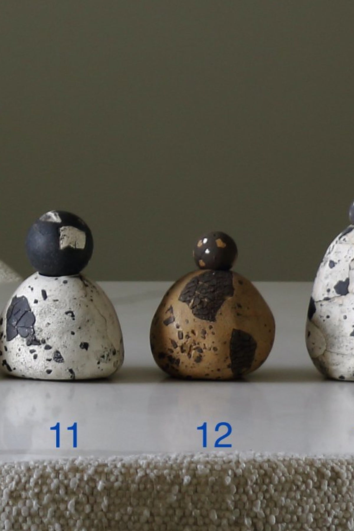 Three small decorative stone-like figurines with speckled tops are arranged on a white surface against a muted taupe background. Two are shown front-on, labeled 11 and 12 in blue text, and a third is partially visible on the right.