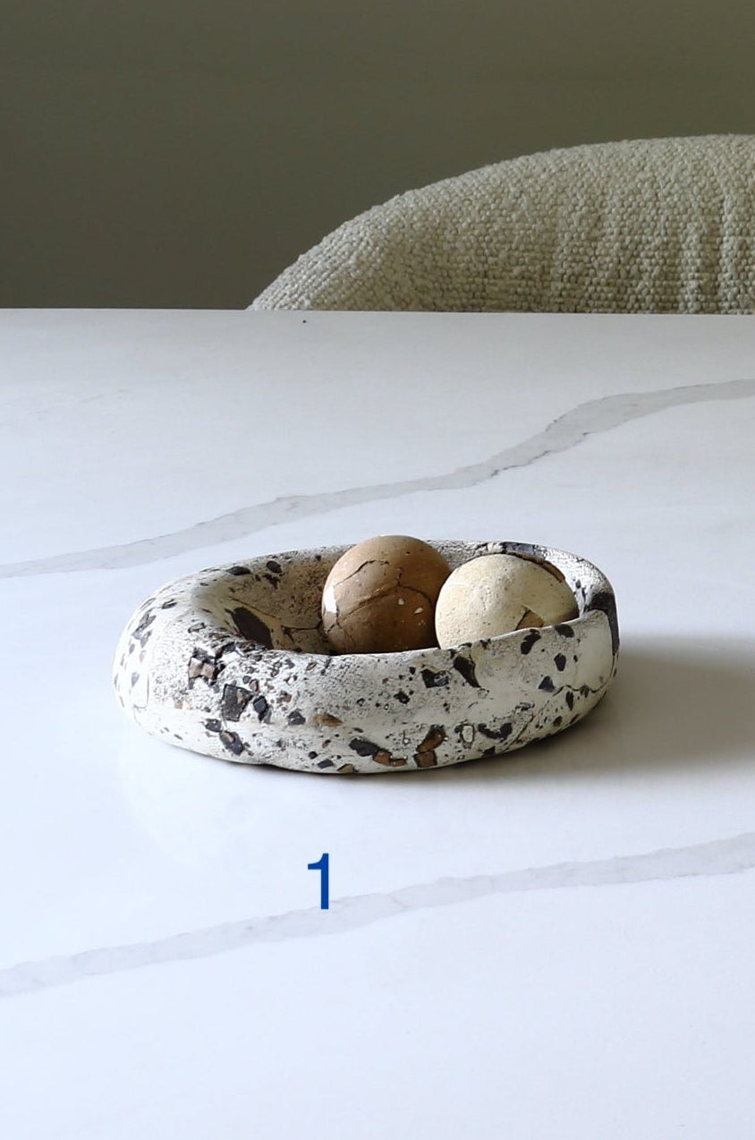 A shallow round ceramic dish with a speckled white, gray, and black pattern, holding two small decorative eggs, set on a white marble table with a textured chair in the background.