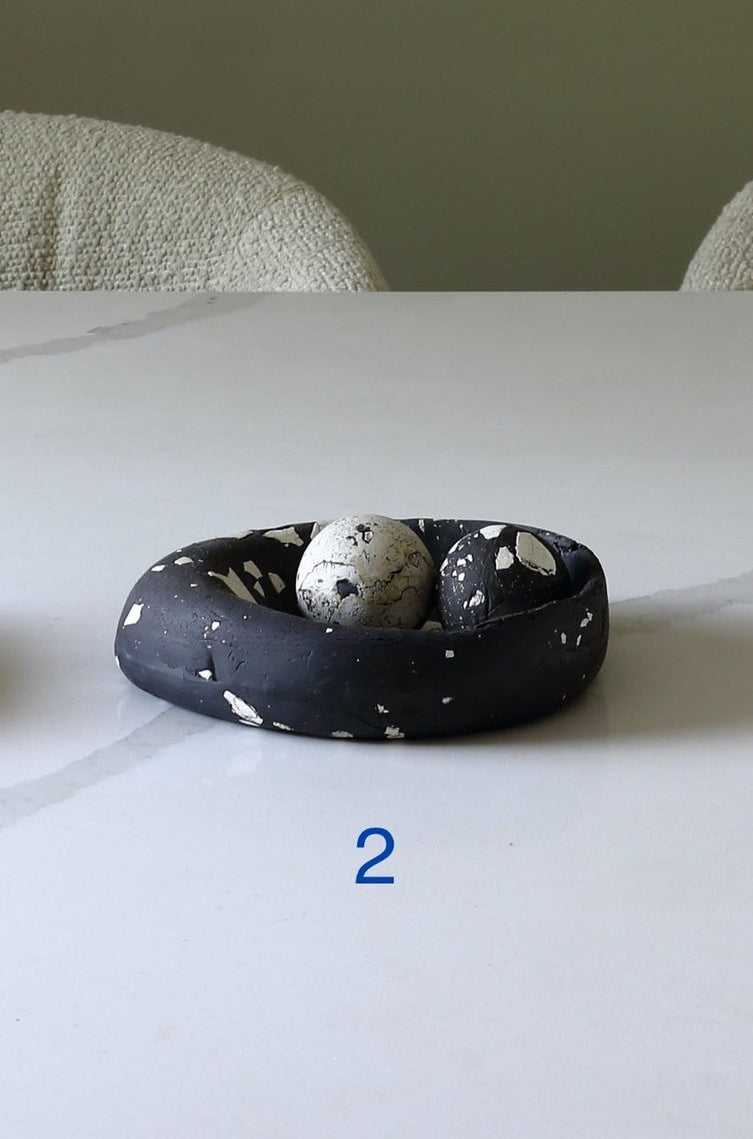 A small black clay nest with white speckles holding a speckled white egg, displayed on a white marble table with two chairs in the background.