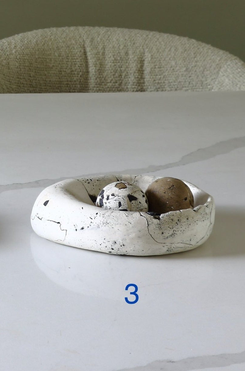 A small white ceramic dish with black speckles sits on a white marble table, holding two decorative eggs. A textured beige chair is visible in the background against a muted green wall.