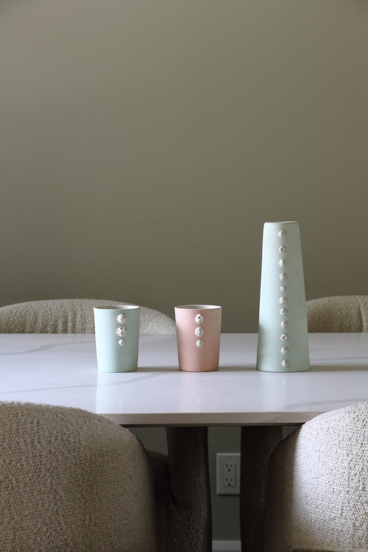 Three decorative ceramic table vases in sage green, dusty pink, and light sage green with white pearl-like accents, arranged on a white table in a dining setting.