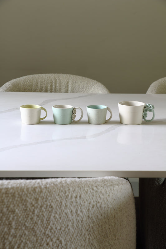 Four ceramic mugs in soft pastel colors lined up on a white table in a minimalist dining setting.