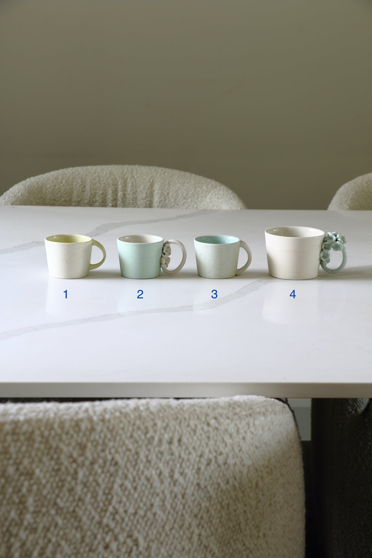 Four ceramic mugs in different pastel colors are lined up on a white table, with blue numbers 1 through 4 printed underneath each mug. The background shows a neutral wall and textured chairs.