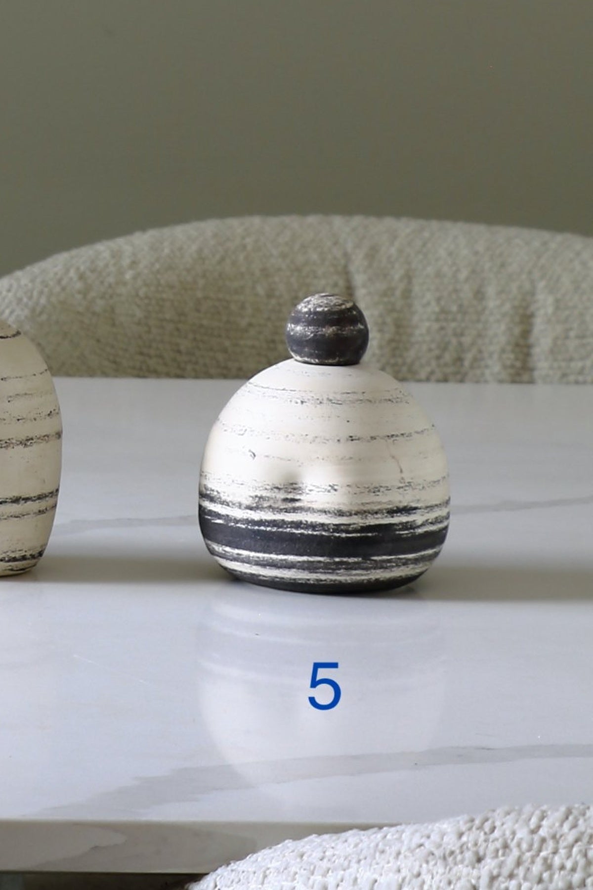A small round ceramic lidded container with a white and black striped pattern and a dark gray spherical lid, displayed on a white marble tabletop with a textured beige chair in the background.