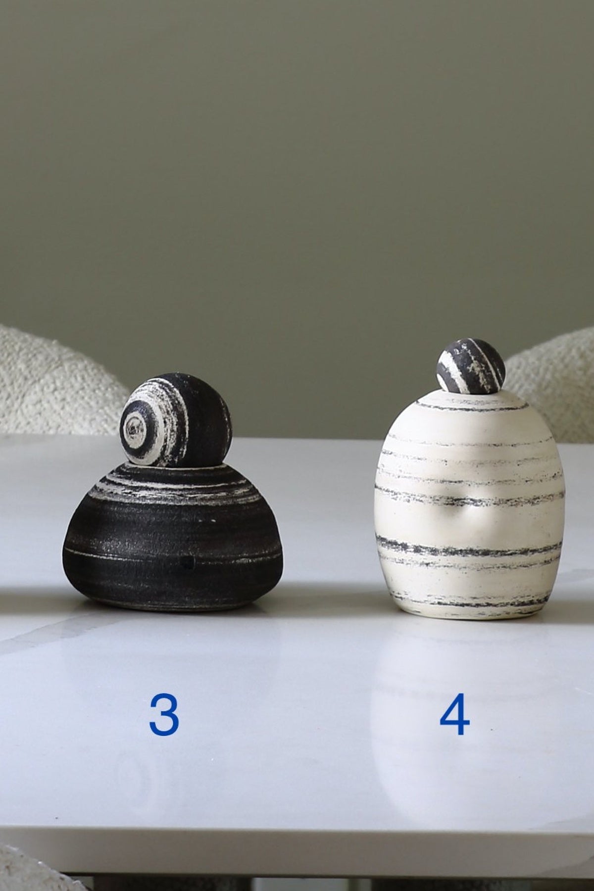 Two small decorative vases or containers with rounded bodies and rounded lids sit on a white tabletop. The left one is glossy black with white spiral bands, and the right one is off-white with dark horizontal stripes.