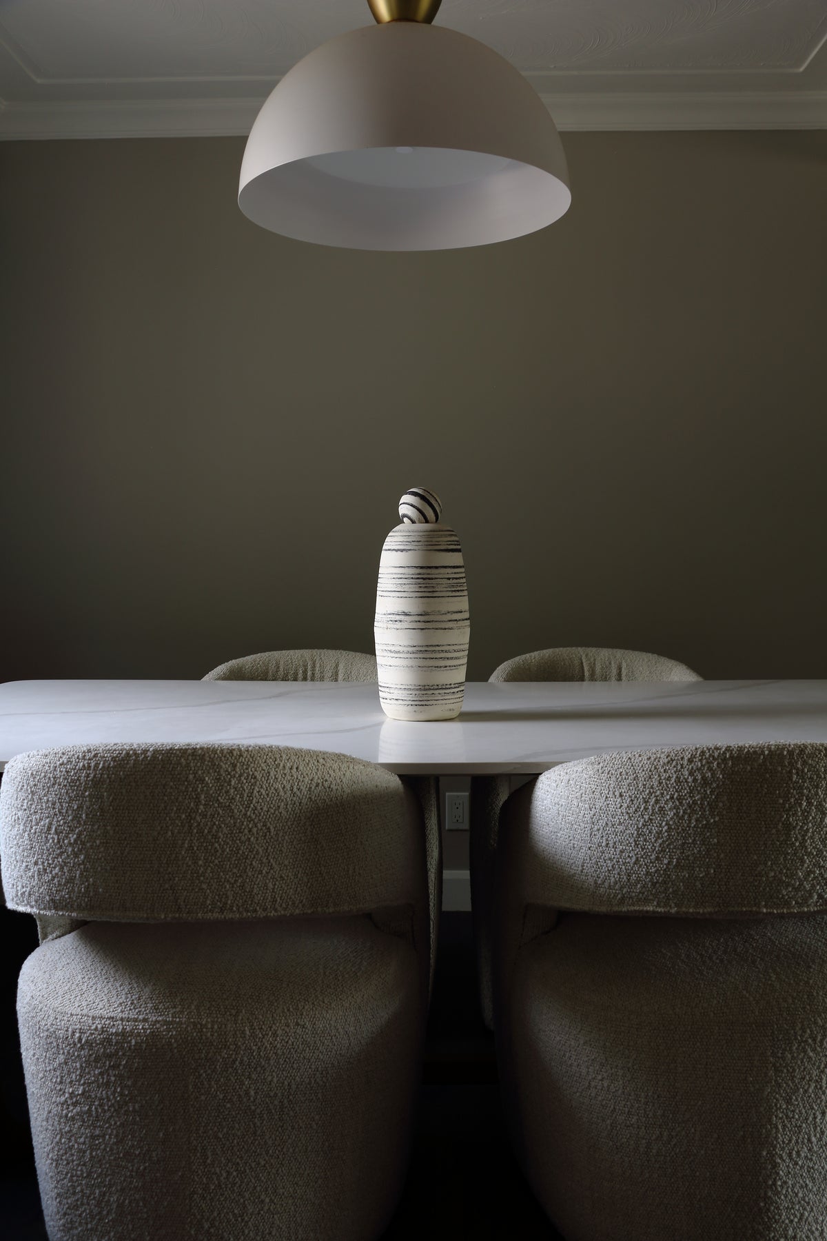 A modern dining room scene featuring a white table, four textured boucle dining chairs in beige, and a central striped ceramic vase. A large white dome pendant light hangs from the ceiling against a dark gray wall.