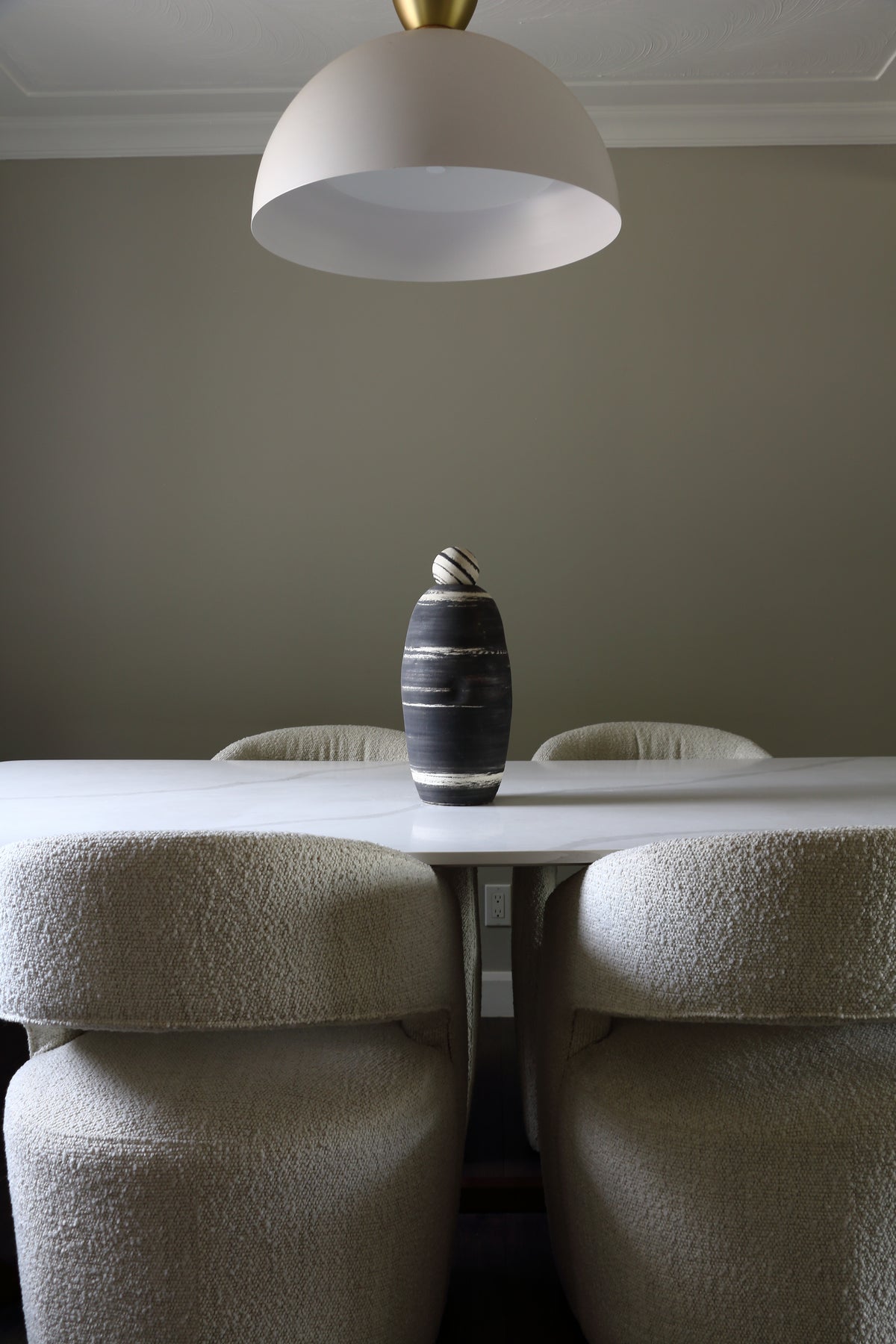 A modern dining room scene featuring a dark textured ceramic vase on a white table, surrounded by four upholstered dining chairs under a large white dome pendant light.