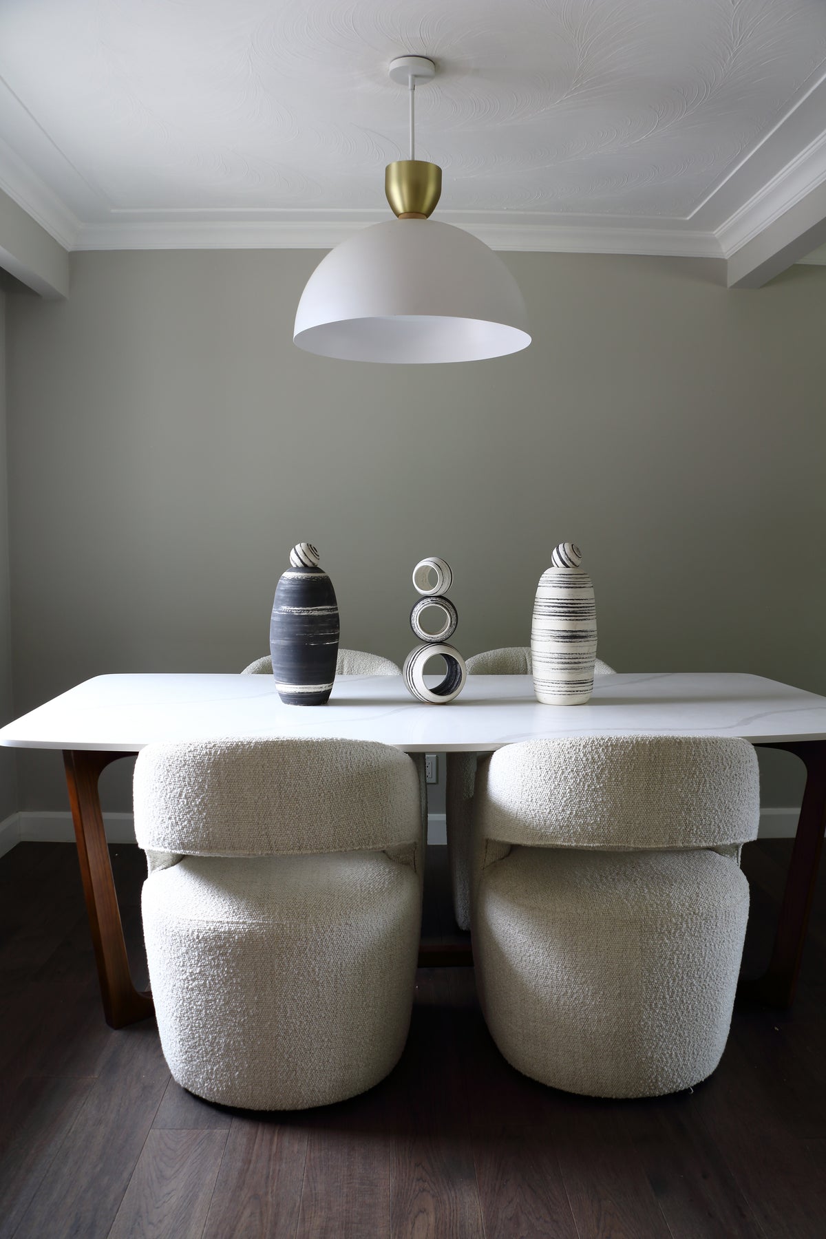 A dining room scene with a white table and four textured cream boucle chairs under a white dome pendant light with a gold cap. Decorative vases sit on the table against a muted sage green wall and dark wood floor.