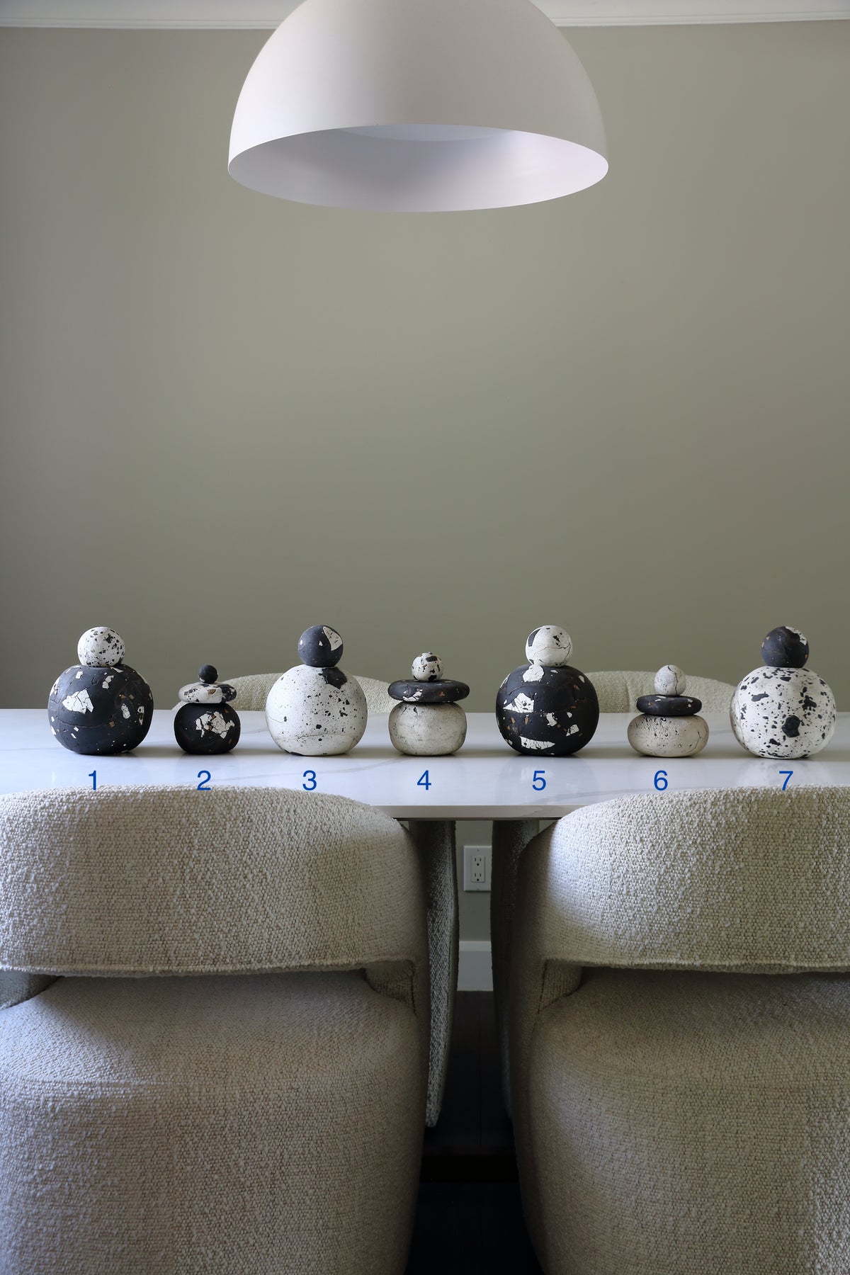 Seven small decorative ceramic duck figurines in black and white speckled patterns are lined up on a white table between two beige boucle armchairs, with blue numbers 1 through 7 printed underneath each duck. A large white dome pendant light hangs from the ceiling above.