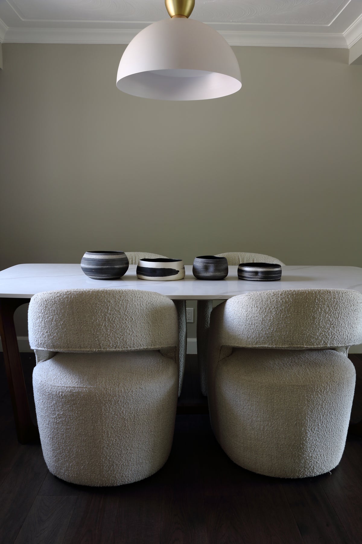 A dining area scene with a white table, two upholstered chairs in a textured cream bouclé fabric, a white dome pendant light with a gold top, and four dark ceramic bowls on the tabletop.