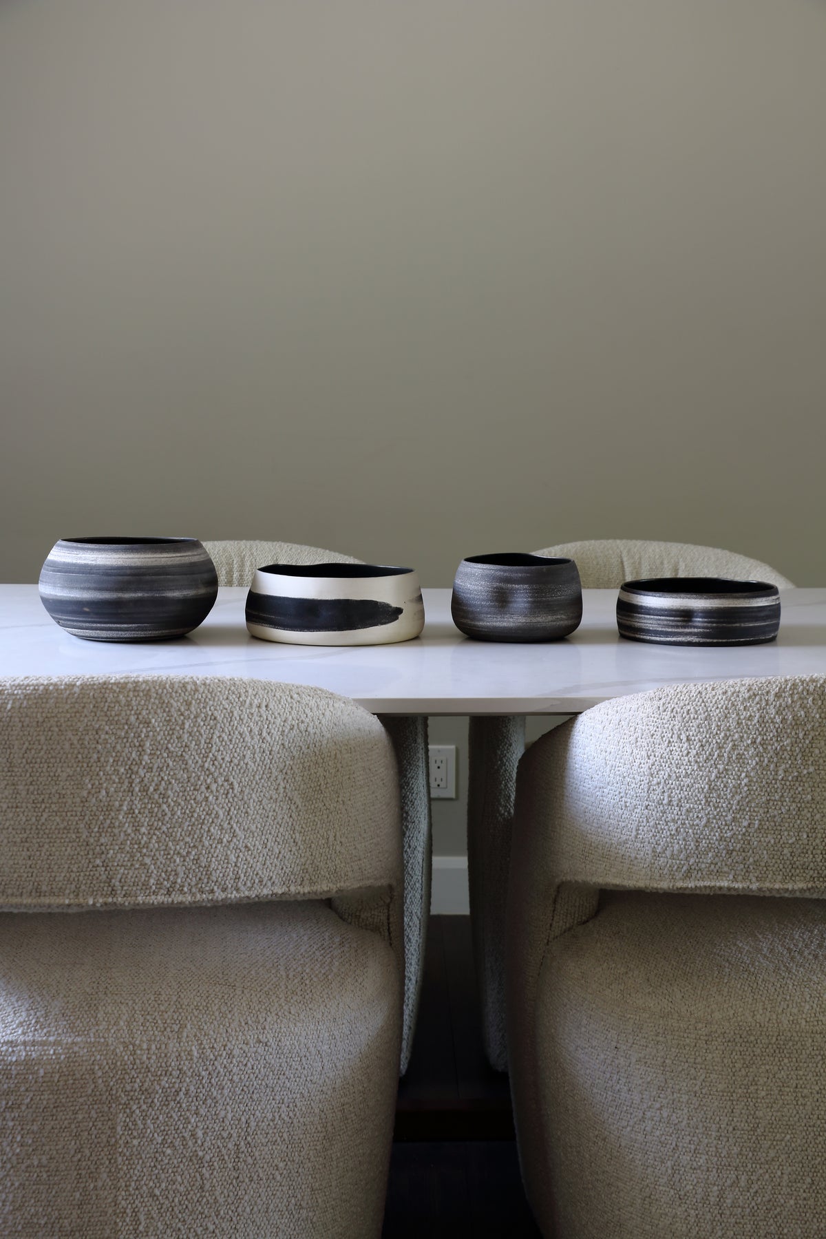 Four round ceramic bowls with black and white marbled patterns arranged on a white table between two upholstered chairs against a neutral beige wall.