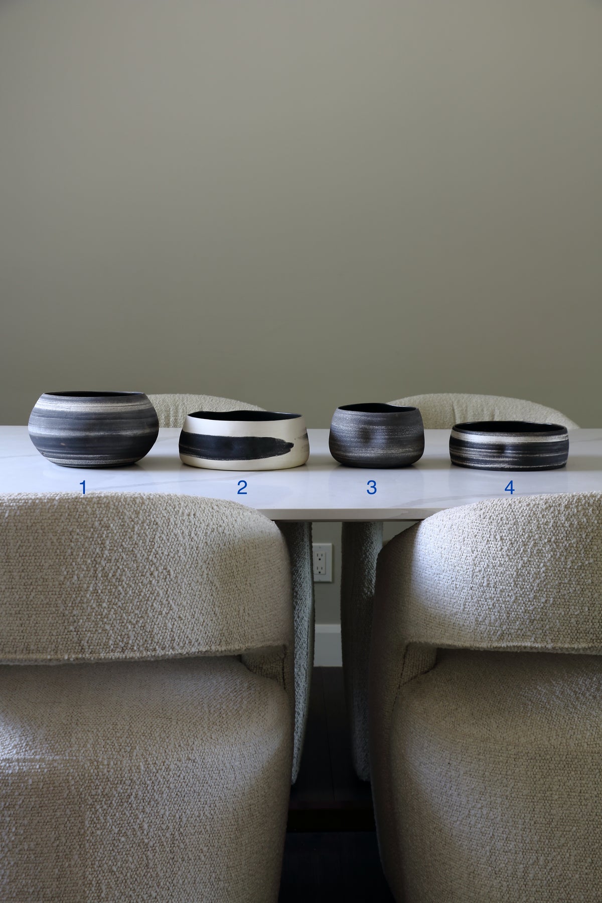 Four round ceramic bowls with black and white marbled patterns arranged on a white table between two upholstered chairs against a neutral beige wall.