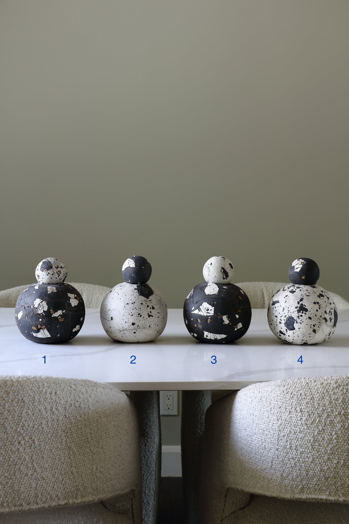 Four decorative ceramic globes with matching lids arranged on a white table in a dining or living room setting, with upholstered chairs visible below and a plain beige wall behind.