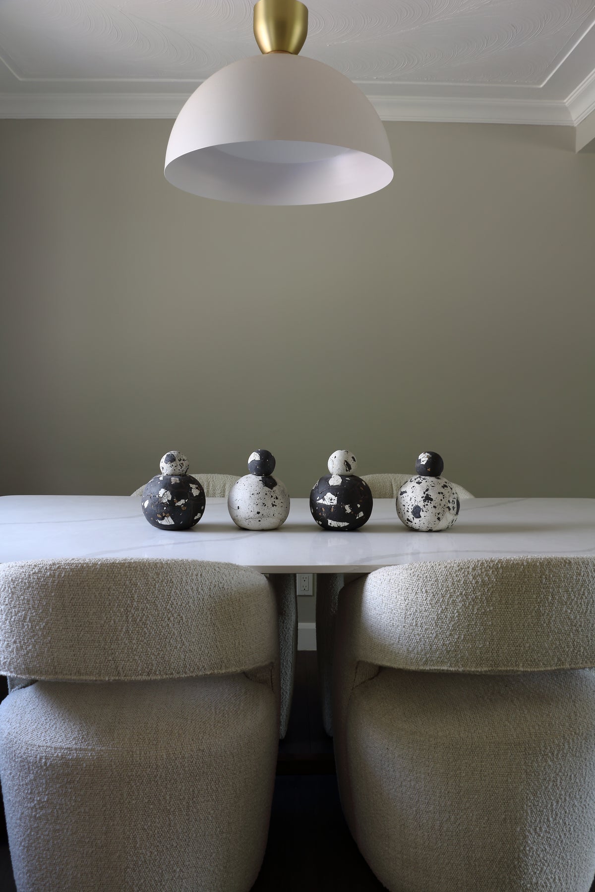 A modern dining room scene featuring a white dome pendant light with a gold finial hanging above a white table. Four decorative ceramic bird figurines in black and white speckled patterns are lined up on the table, with two matching textured beige upholstered dining chairs in the foreground.