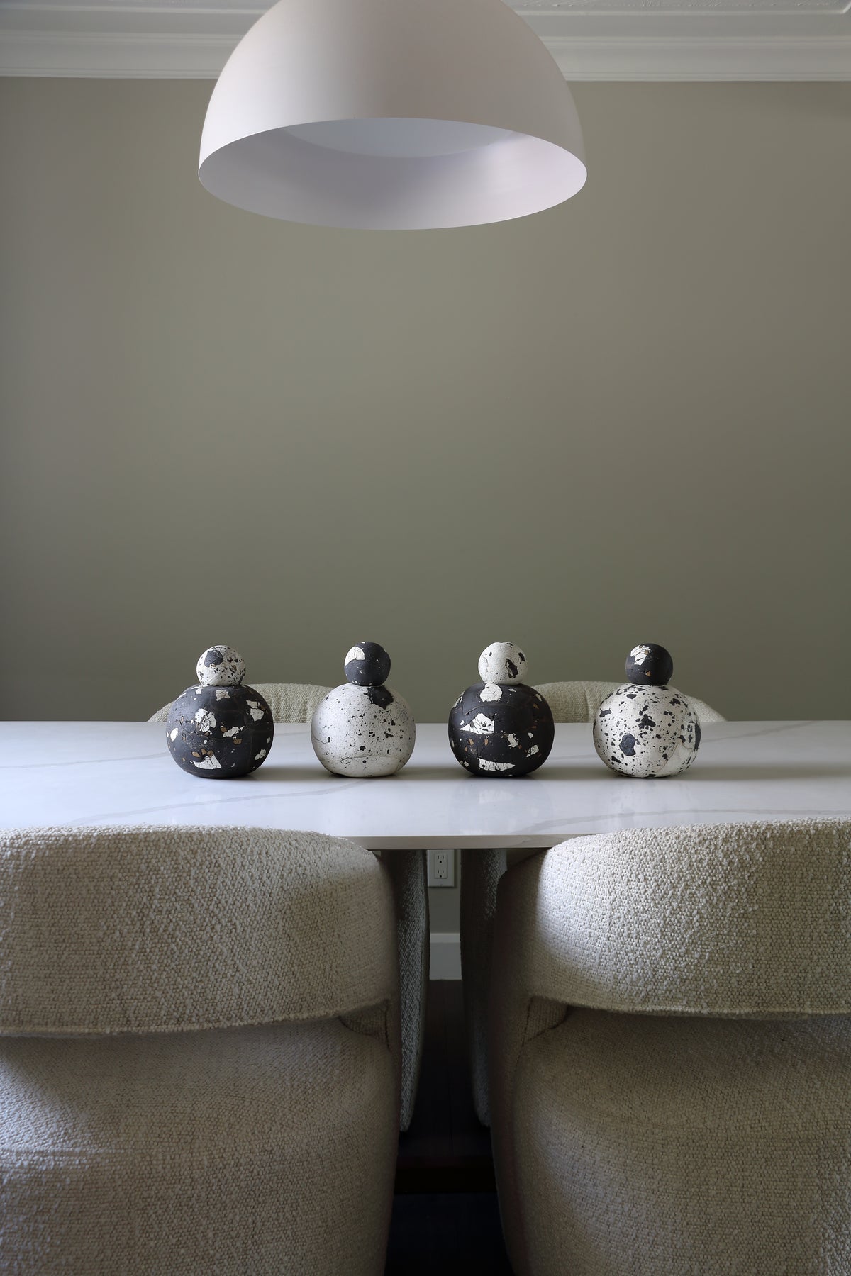 Four decorative duck-shaped sculptures in black and white speckled ceramic, arranged on a white dining table between two upholstered chairs under a white dome pendant light.