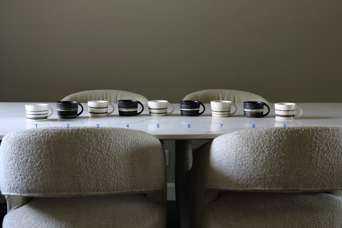 Nine ceramic mugs in various color combinations of black, white, and beige stripes, arranged in a row on a table in front of three upholstered chairs.