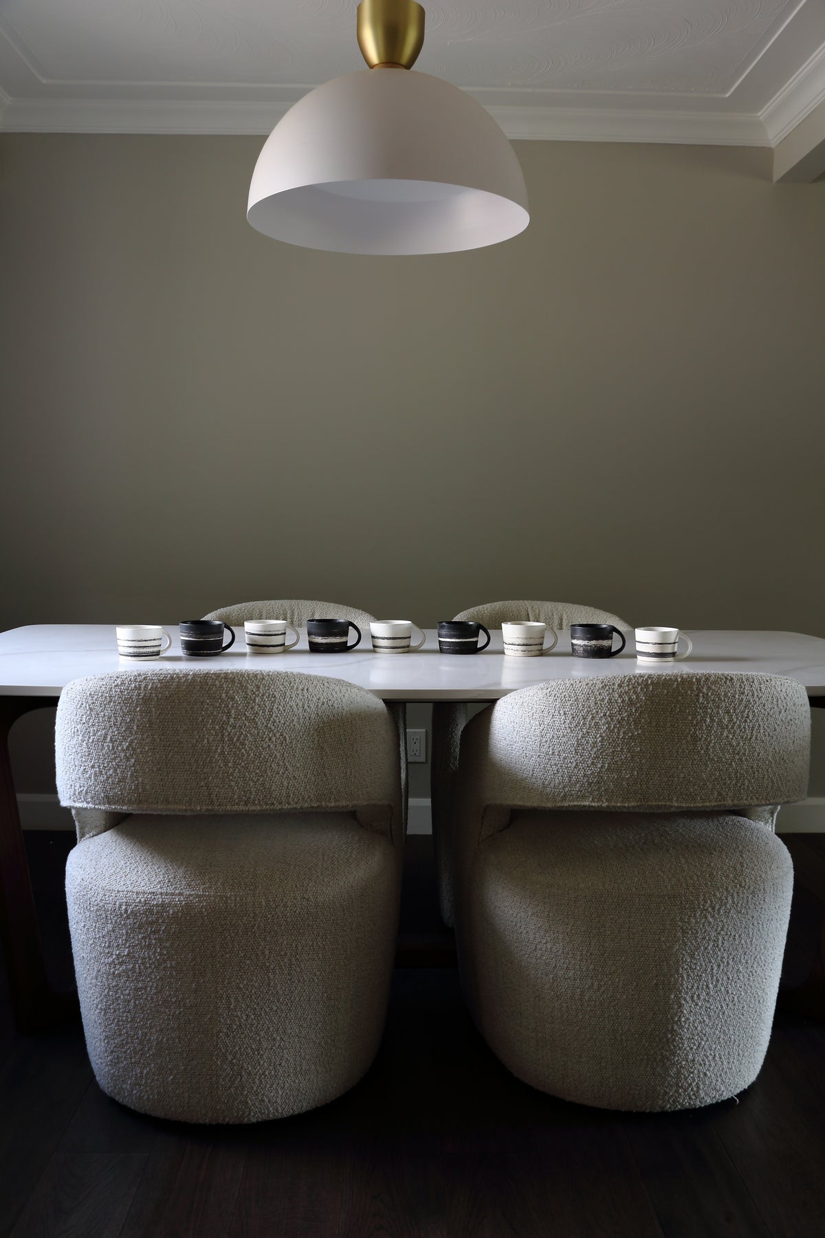 A dining room scene with a long table set with small white dishes and mugs, four upholstered round chairs in a textured cream bouclé fabric, and a large white dome pendant light hanging above against a muted gray wall.