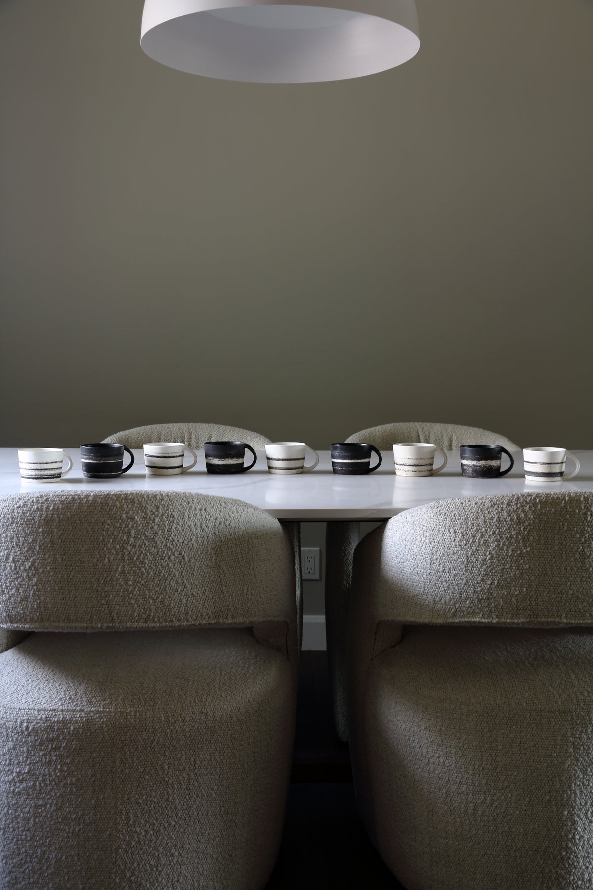 A dining table set with multiple black and white ceramic mugs and saucers, flanked by two upholstered chairs in a minimalist room with a dark gray wall and a white pendant light overhead.
