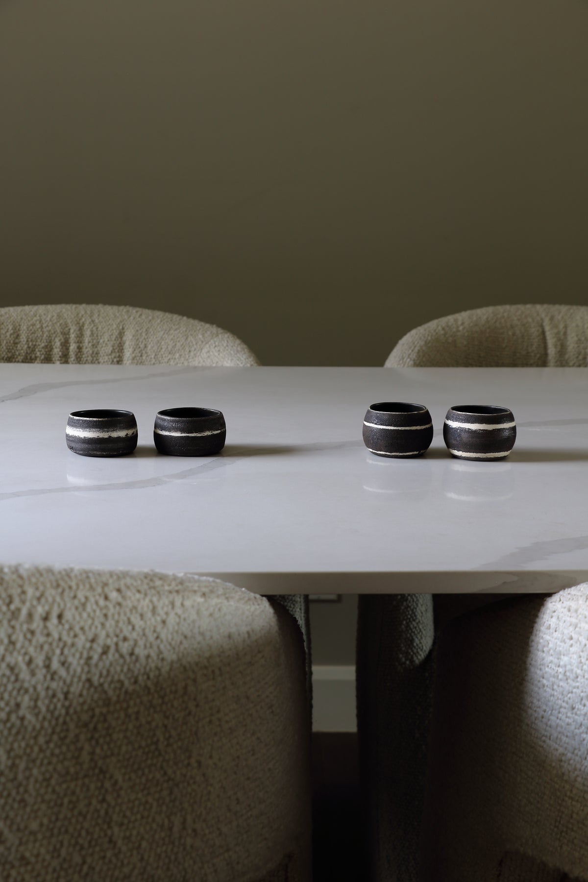 Four black ceramic rings with metallic silver bands, arranged on a white marble dining table surrounded by beige upholstered chairs.