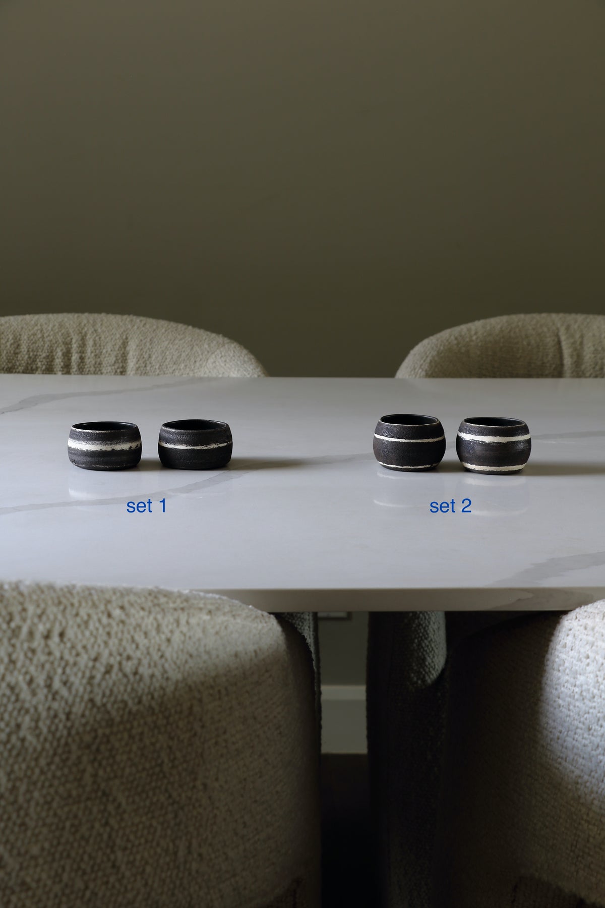 Four black ceramic table rings with metallic silver bands are arranged on a white marble dining table, split into two sets of two labeled 'set 1' and 'set 2' in blue text. The background features neutral beige upholstered dining chairs against a dark gray wall.