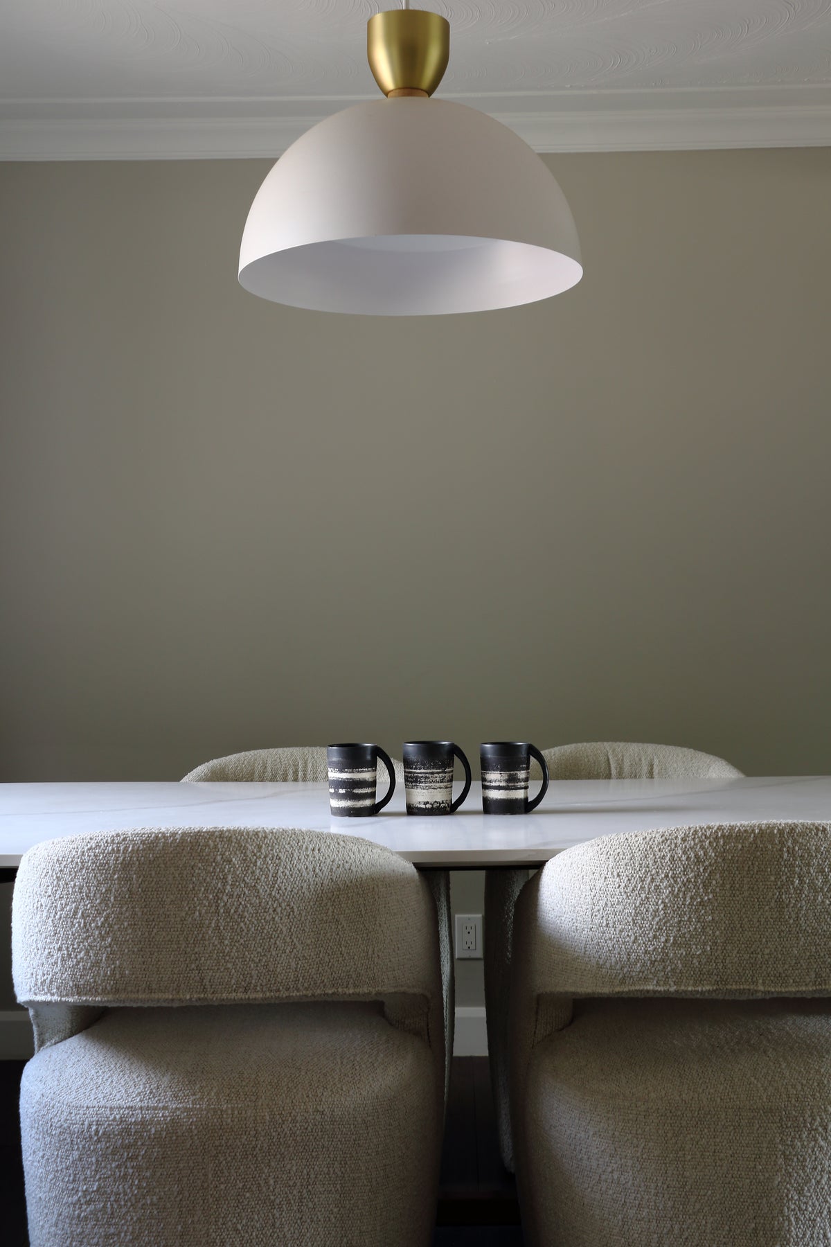 A modern dining room scene featuring a large white dome pendant light with a gold ceiling mount hanging over a white table with textured beige boucle chairs. Three black mugs sit on the table against a neutral gray wall.