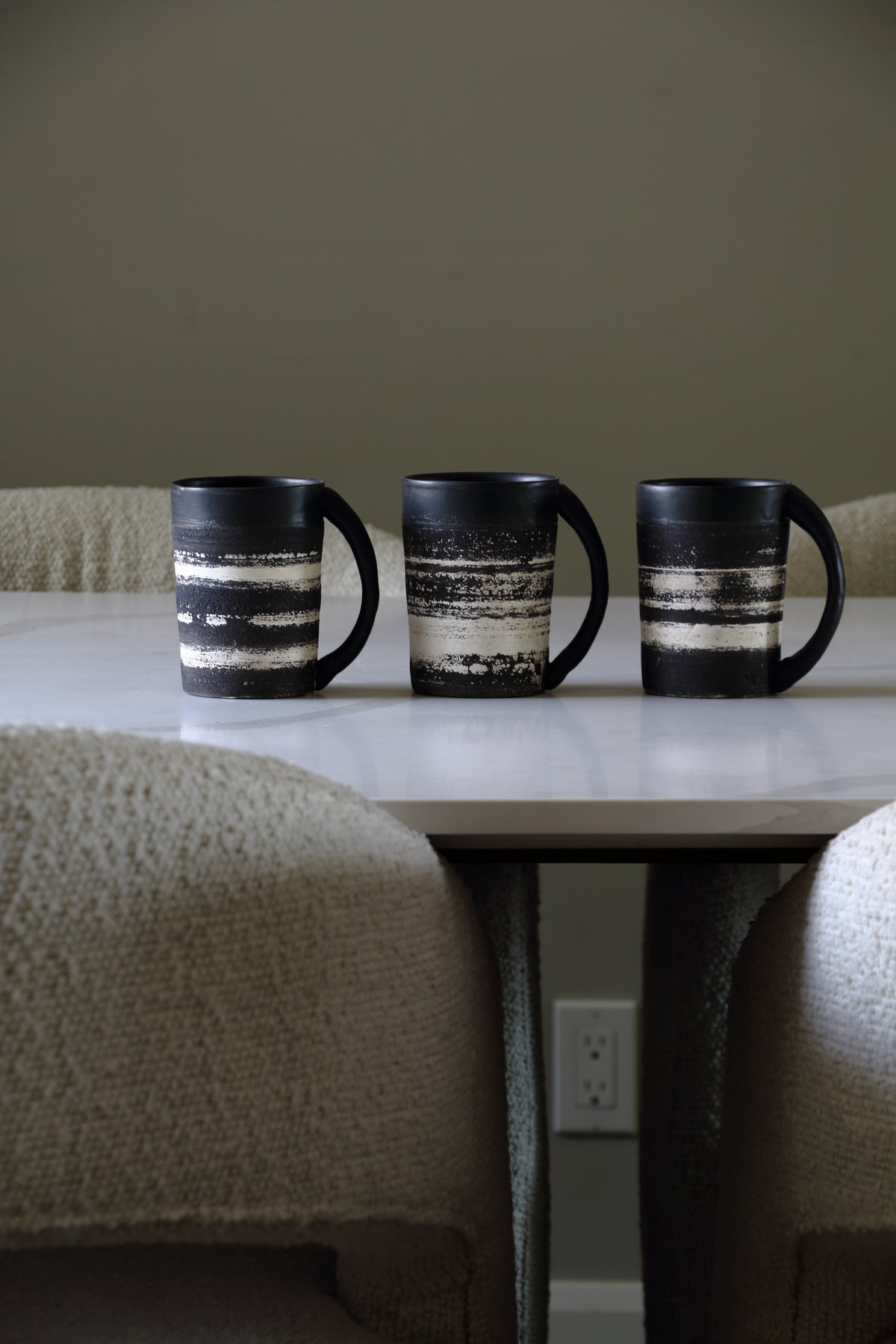 Three black ceramic mugs with horizontal distressed white stripes are lined up on a white table in a dining area, with blue numbers 1, 2, and 3 printed below each mug.