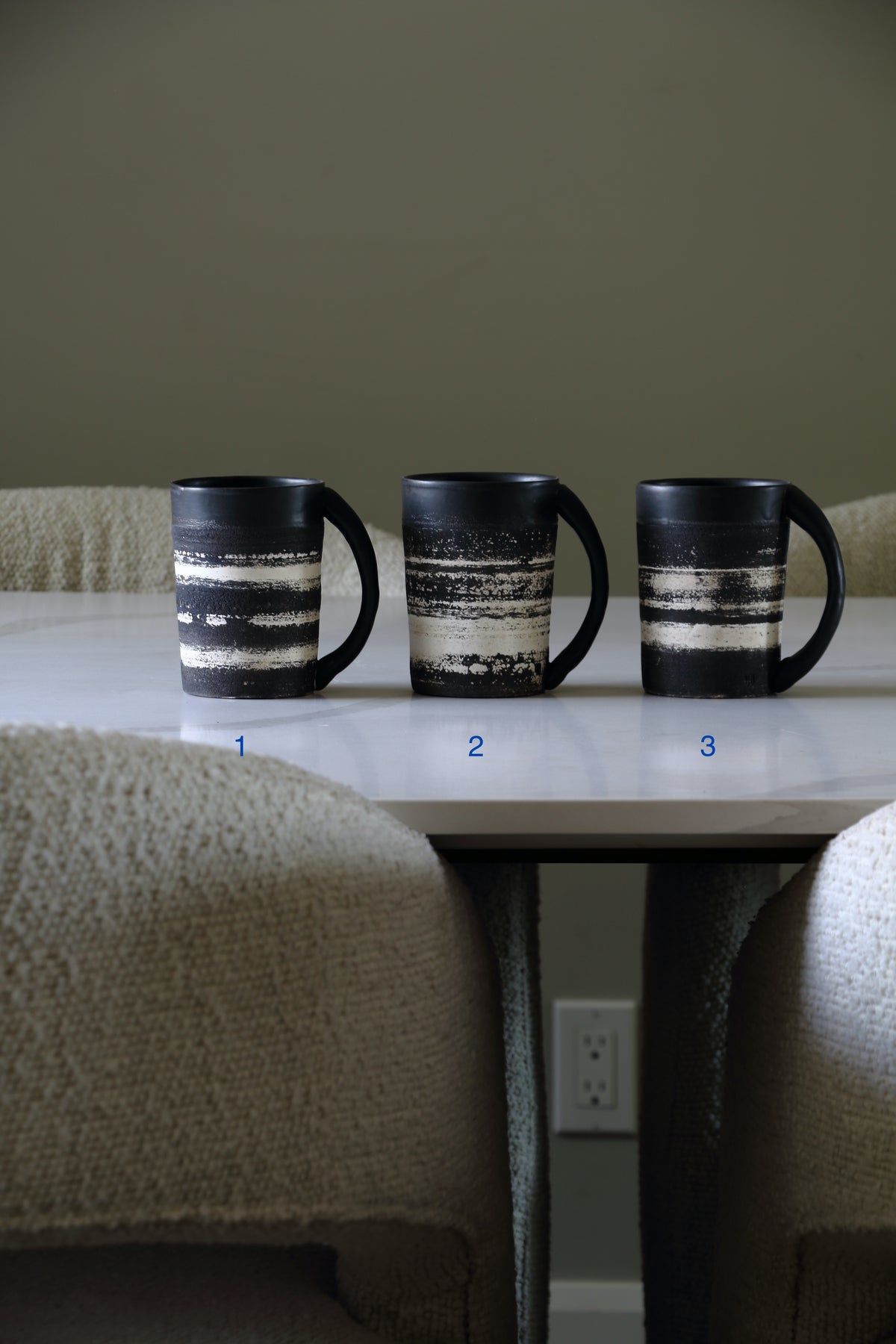 Three black ceramic mugs with horizontal distressed white stripes are lined up on a white table in a dining area, with blue numbers 1, 2, and 3 printed below each mug.