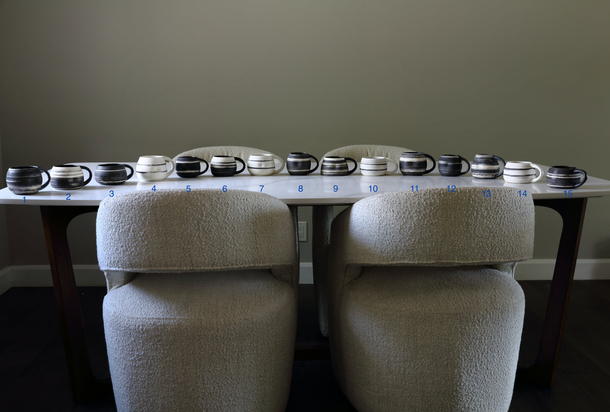 A row of ceramic mugs in various colors and patterns arranged on a white table, with two upholstered chairs in front. Small blue numbers are printed underneath the mugs, likely indicating color or style options.