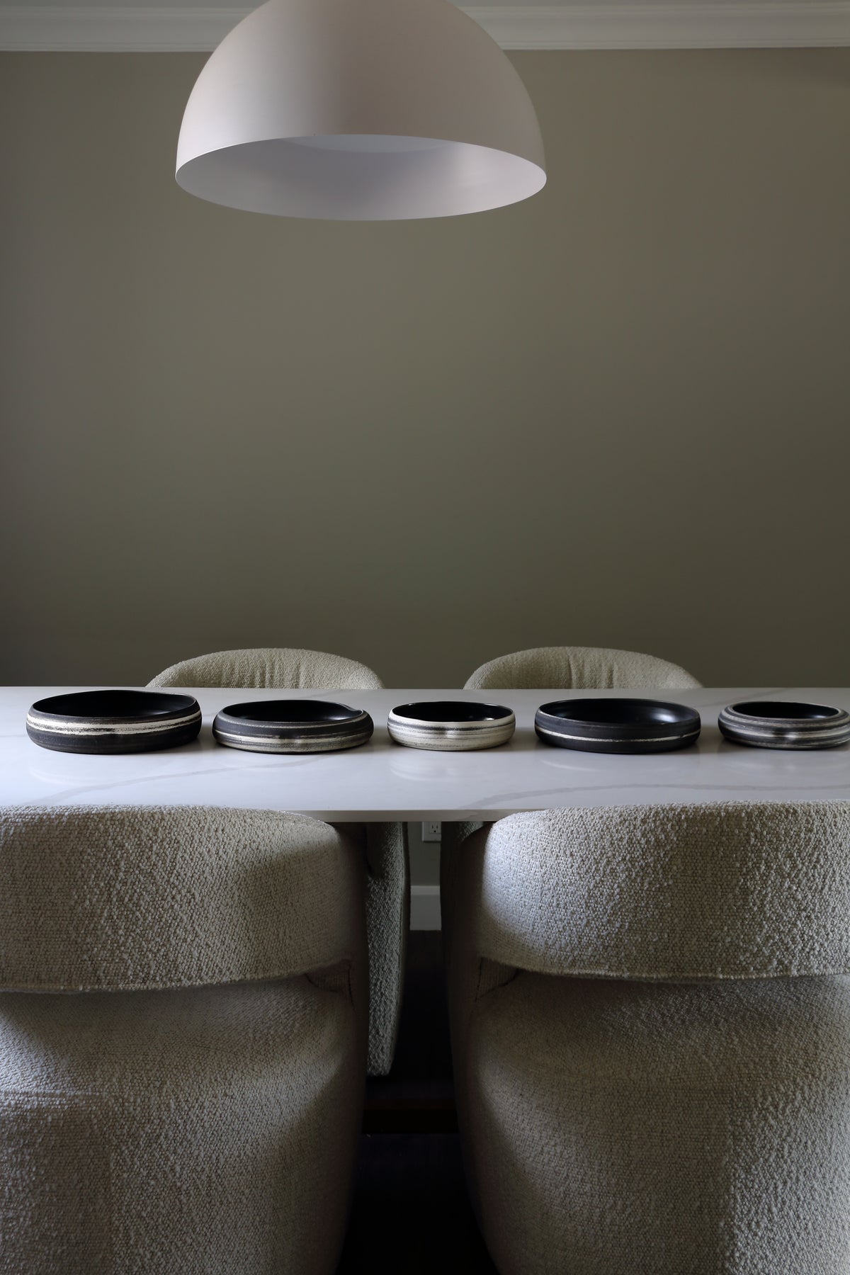 Six round black and silver coasters arranged in a row on a white marble dining table, with four upholstered beige chairs underneath and a large white dome pendant light hanging above against a muted gray-green wall.