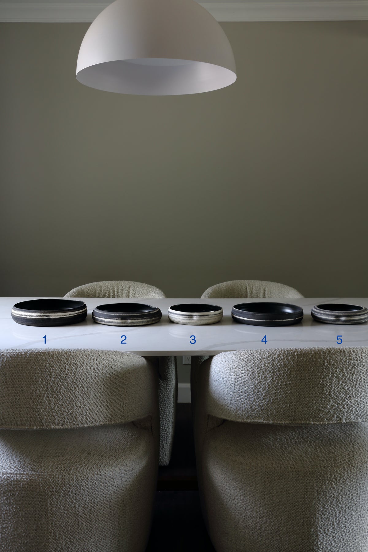 Five circular black and silver metallic rings or plates arranged in a row on a white table, with blue numbers 1 through 5 underneath. The scene is framed by two upholstered beige chairs and a large white dome pendant light against a muted gray-green wall.