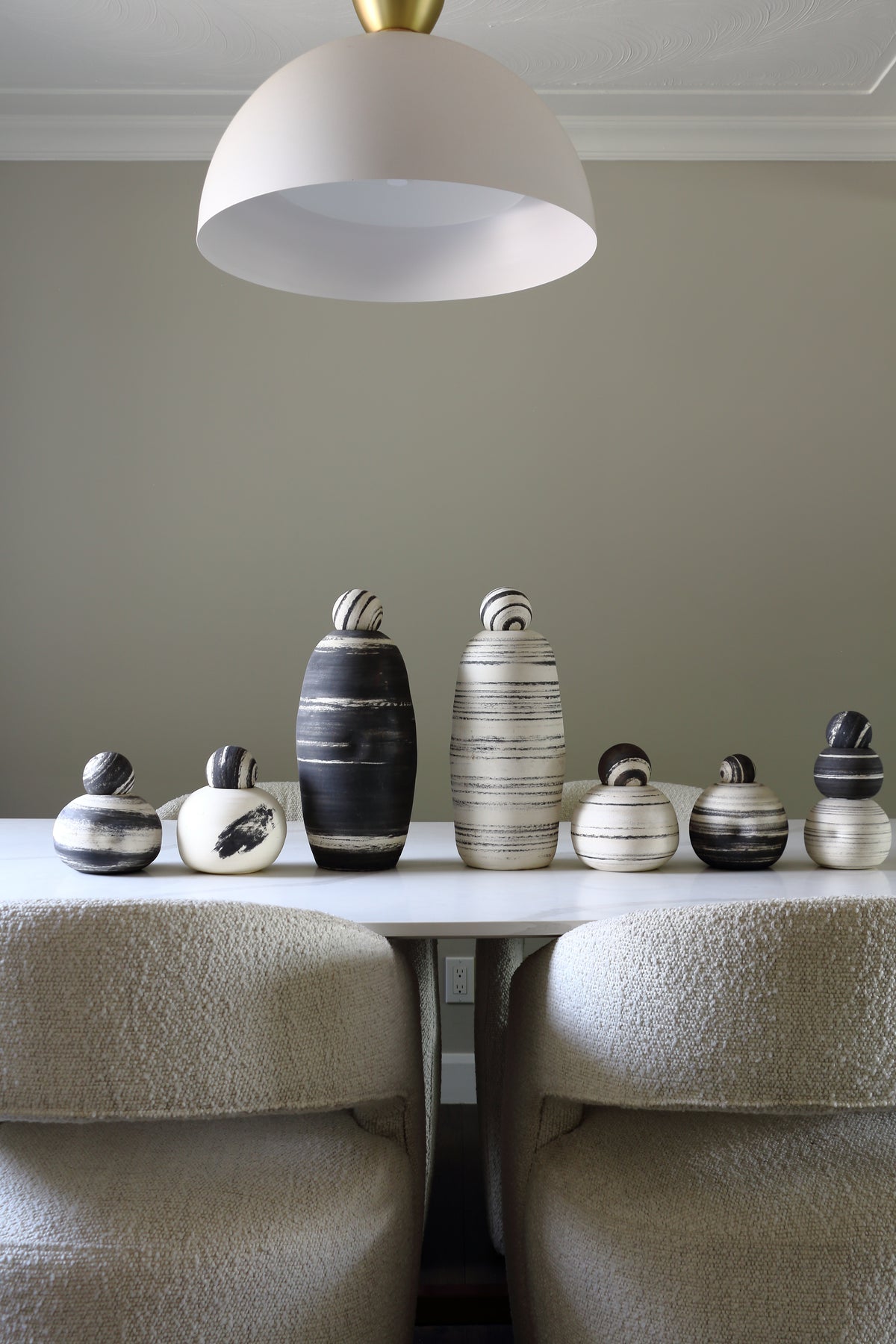 A collection of modern ceramic vases with organic, rounded shapes in black, white, and gray tones, displayed on a white table between two textured beige chairs under a white dome pendant light.