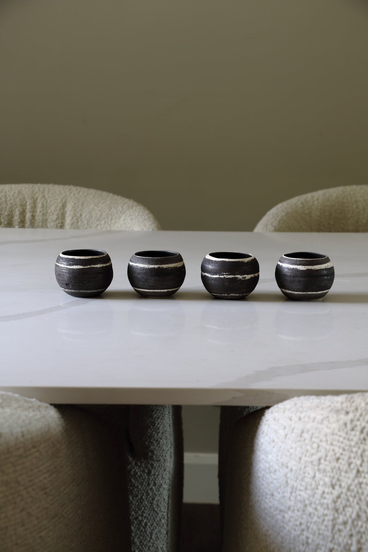 Four small round black ceramic cups with white horizontal stripes lined up on a white marble dining table surrounded by textured beige chairs.