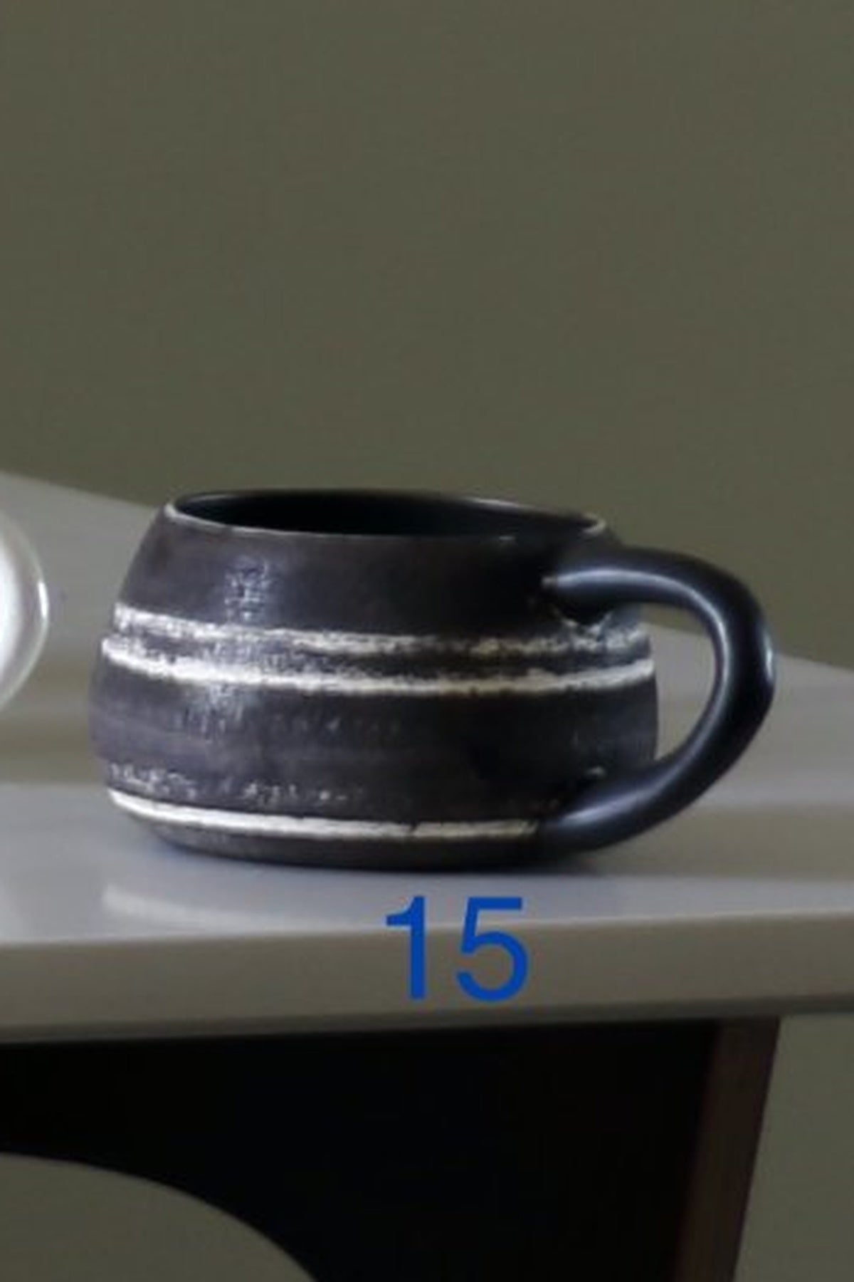 A dark ceramic mug with horizontal white textured stripes and a glossy black handle, sitting on a white tabletop with a blue '15' overlay.