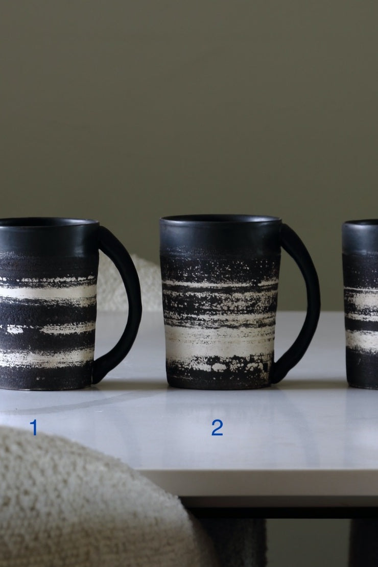 Three ceramic mugs with black handles and a distressed black-and-cream horizontal striped pattern, displayed on a white table against a muted olive background.