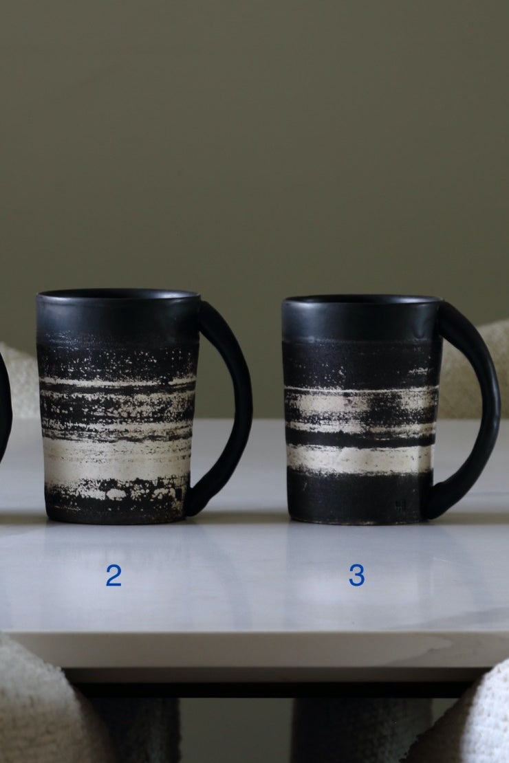 Two ceramic mugs with black handles and bodies, decorated with horizontal distressed stripes in cream and black, displayed on a white table against a muted olive background.