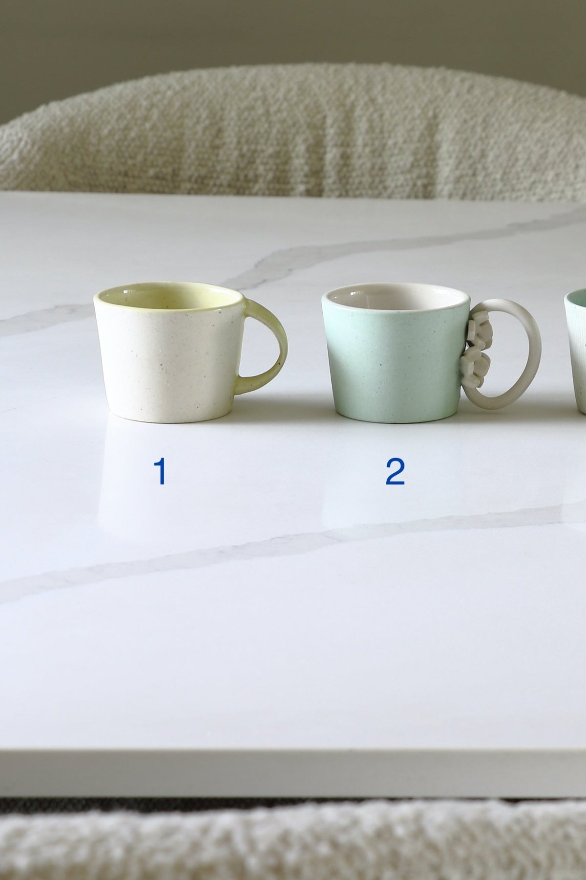Two ceramic mugs on a white marble table, labeled 1 and 2. Mug 1 is a matte off-white with a pale yellow interior, and mug 2 is a pale mint green with a white interior and a small floral accent on the handle.