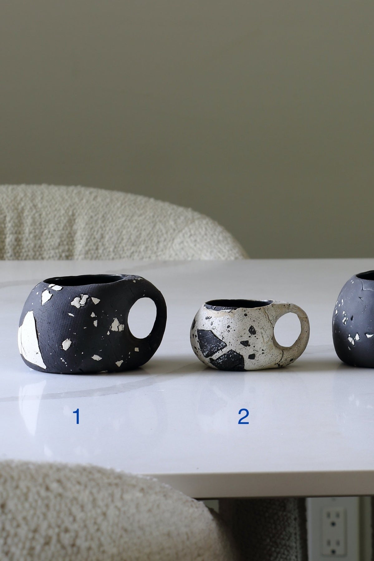 Three handmade ceramic cups with terrazzo-style speckles are lined up on a white table. Cup 1 is black with white speckles, cup 2 is white with black speckles, and cup 3 is partially visible on the right.