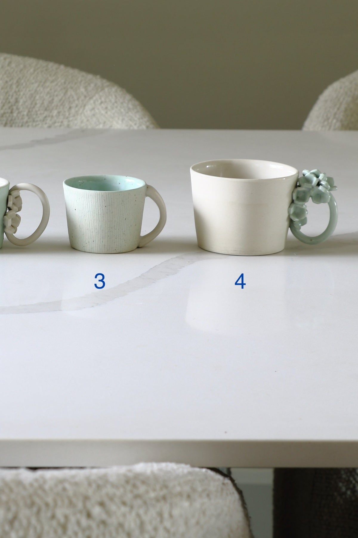 Two ceramic mugs displayed on a white table in a real indoor setting with chairs visible in the background. The left mug is off-white with a pale mint interior, and the right mug is off-white with a pale mint decorative handle.