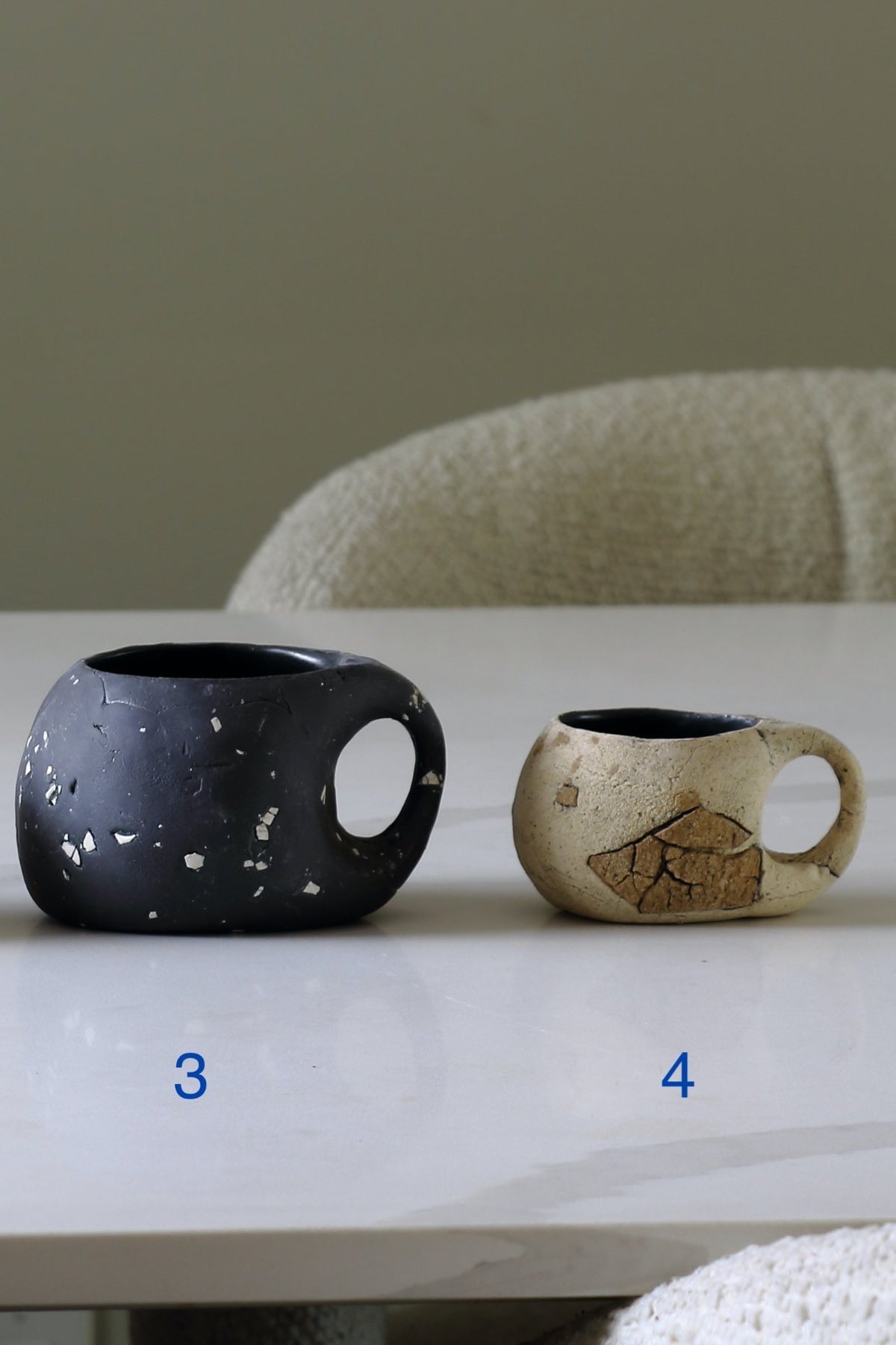 Two ceramic mugs with loop handles sit on a white table. The mug on the left is matte black with white speckles, and the mug on the right is a warm beige with a brown leaf design and dark interior.