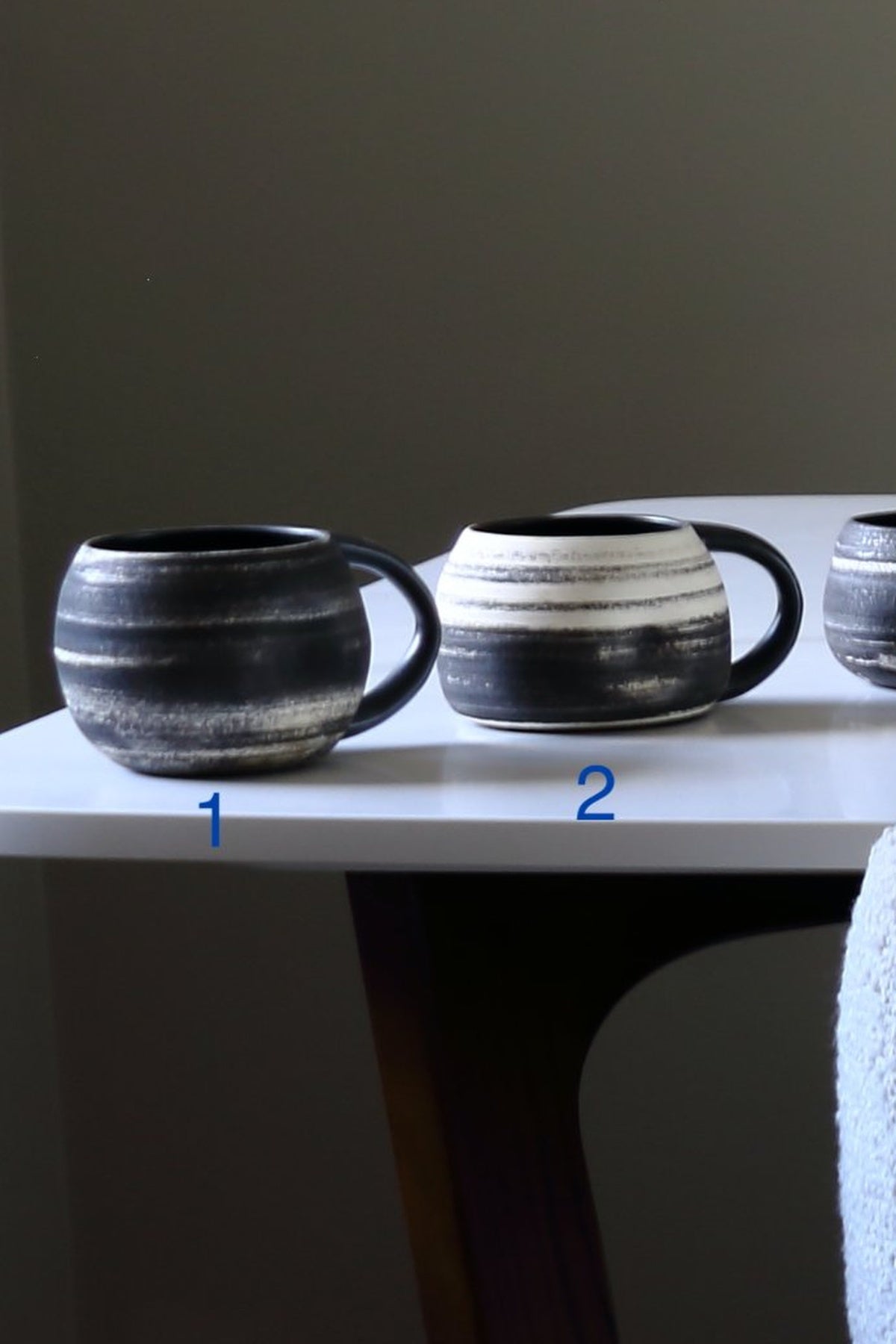 Three handmade ceramic mugs with textured black and white horizontal stripes displayed on a white table. The mugs are numbered 1, 2, and 3 with blue numbers overlaid on the table surface.
