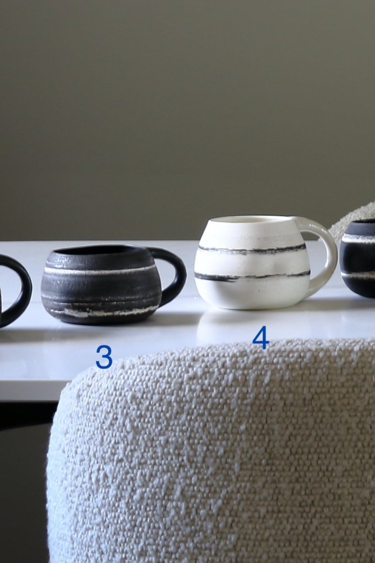 Two ceramic mugs displayed on a white table against a neutral gray wall, with a textured beige chair in the foreground. One mug is matte black with white horizontal stripes, and the other is white with black horizontal stripes.