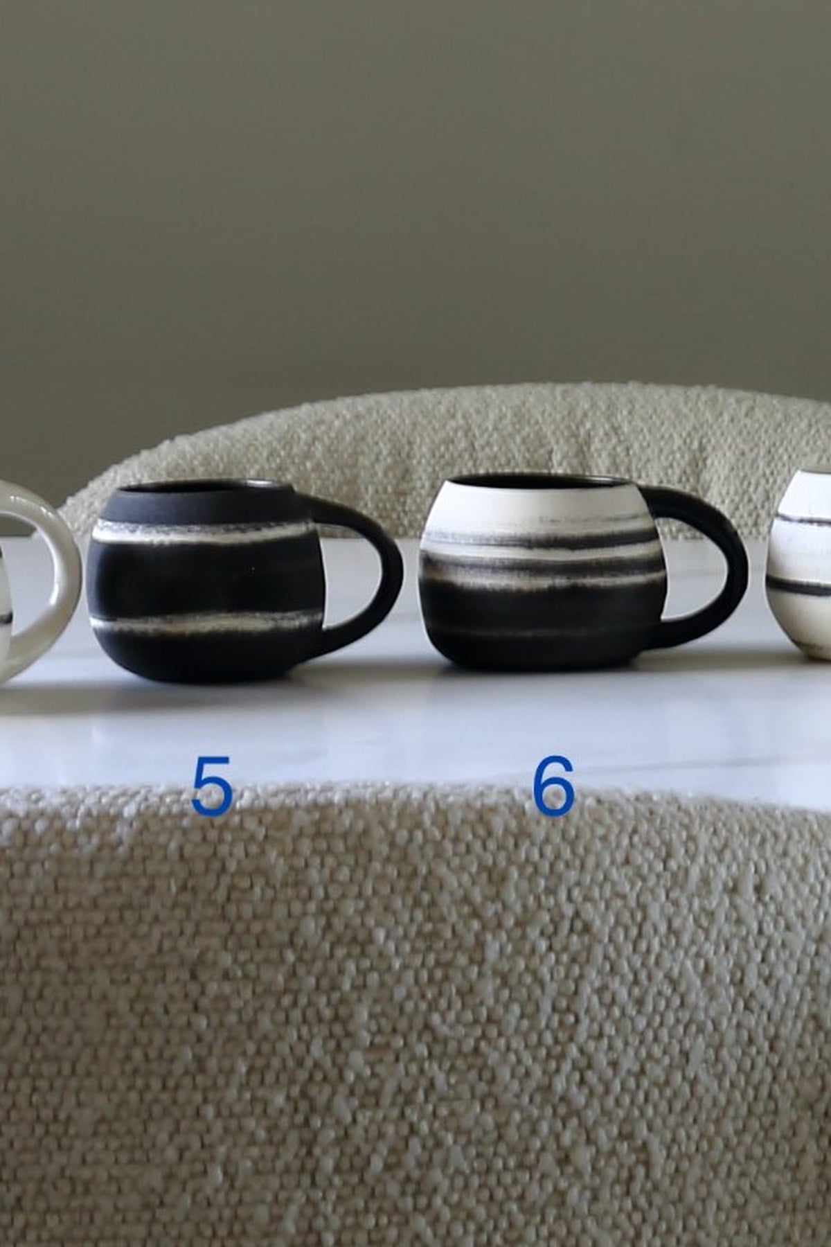 Three ceramic mugs with black and white horizontal stripes sit on a white surface in front of a textured beige fabric backdrop. Blue numbers 5 and 6 are overlaid near the mugs.