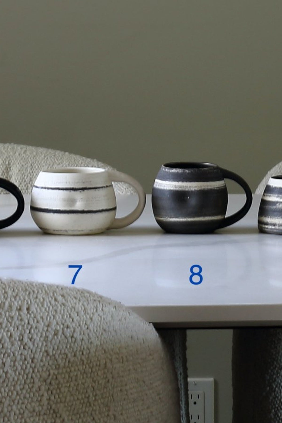 Two ceramic mugs with horizontal stripes displayed on a white table, with blue numbers 7 and 8 overlaid near the base of each mug.