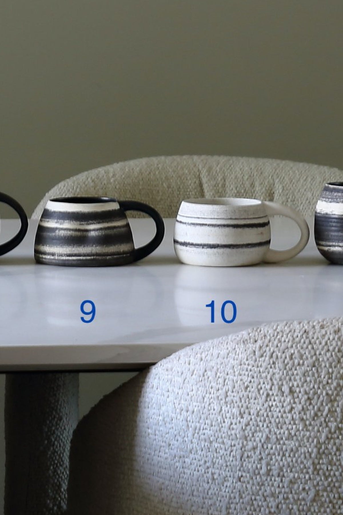 Two ceramic mugs with horizontal stripes are displayed on a white table in front of a textured chair. The left mug is black and white, and the right mug is white and black.
