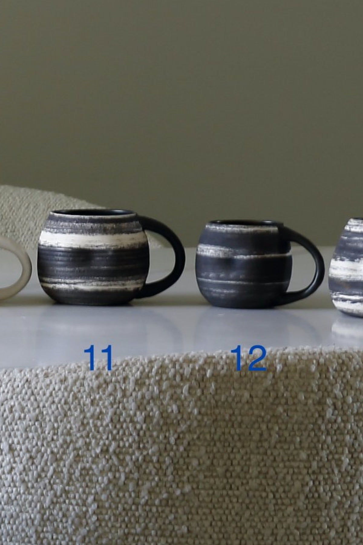 Two ceramic mugs with a black and white horizontal striped glaze are displayed on a white surface. The left mug has the blue number '11' printed below it, and the right mug has '12' printed below it.